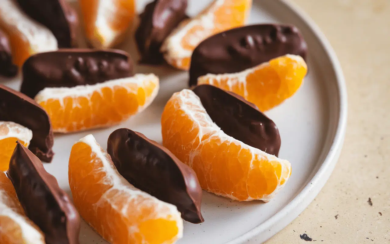 Segments d'orange enrobés de chocolat noir, disposés sur une assiette beige. Savoureux mélange de fruits et de chocolat.