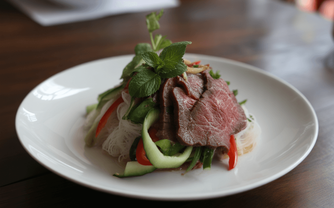 Plat de nouilles avec tranches de boeuf, légumes colorés (poivrons, concombre) et garniture de menthe fraîche. Présenté dans une assiette blanche.