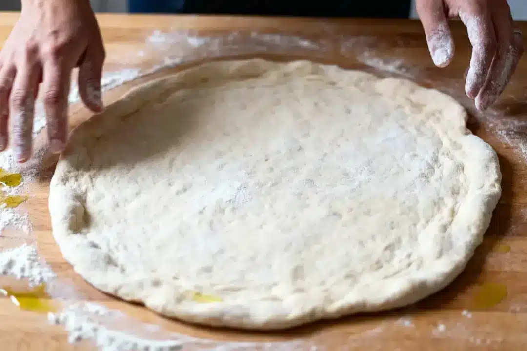 Deux mains façonnant une pâte à pizza sur une surface enfarinée.