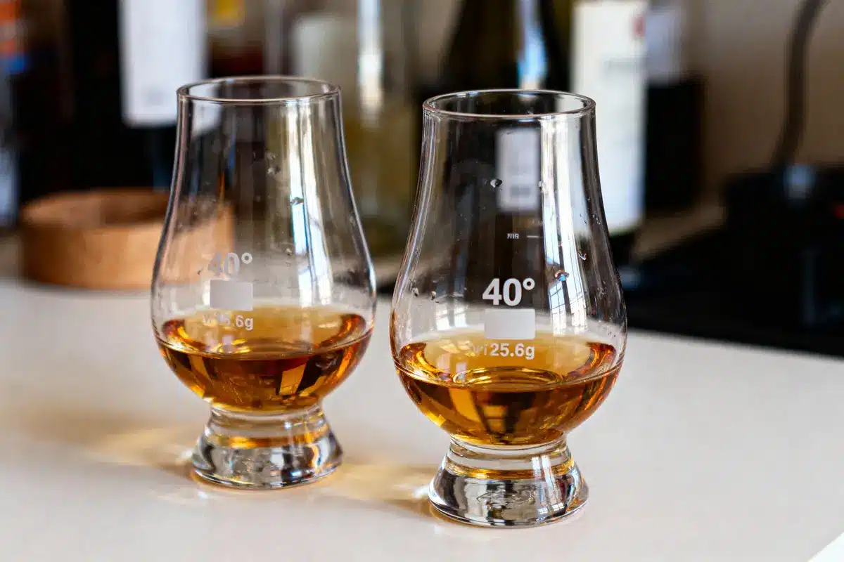 Deux verres remplis de whisky sur une table, avec marquage "40°" et "25.6g", bouteilles floues en arrière-plan.