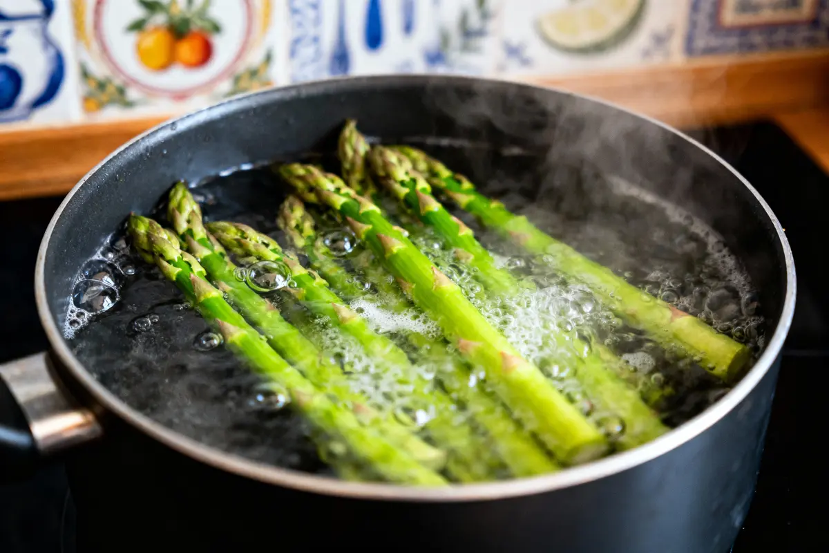 Asperges vertes bouillies dans une casserole noire sur cuisinière, carrelage coloré en arrière-plan.