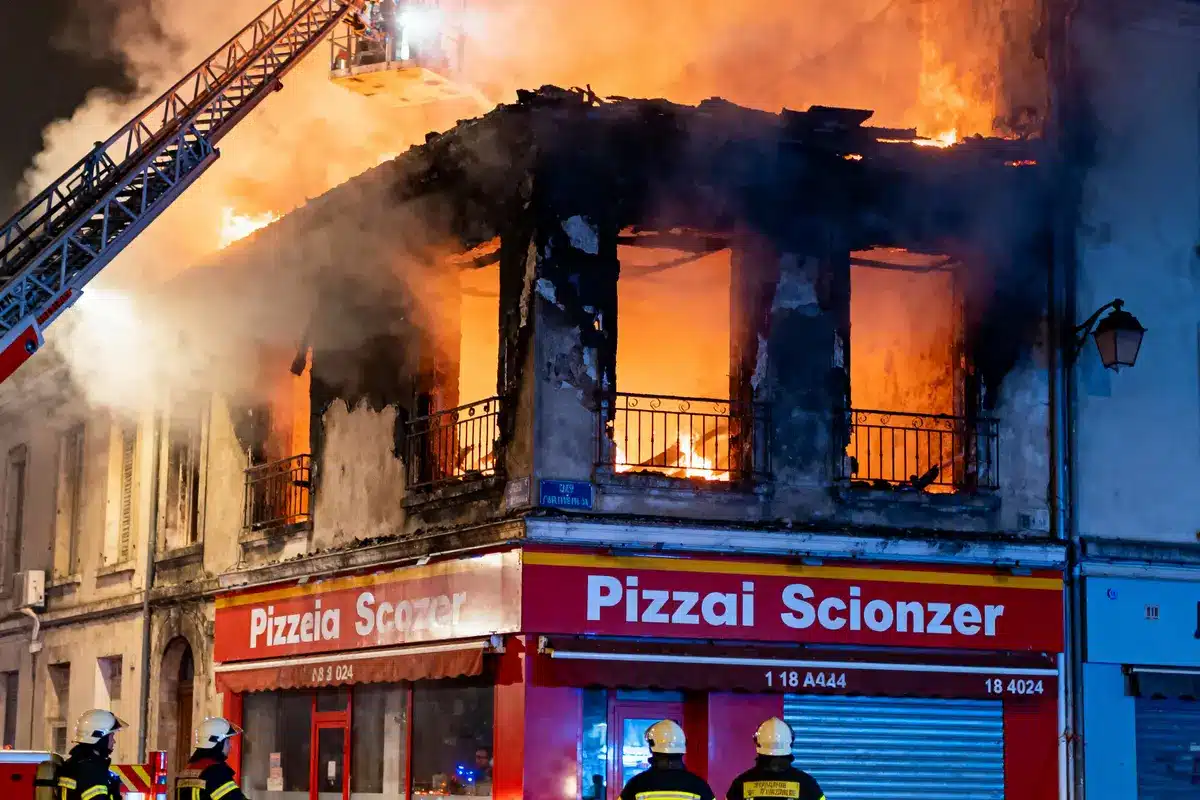 Incendie à l'étage d'une pizzeria. Pompiers en intervention, flammes visibles, échelle déployée, nuit.
