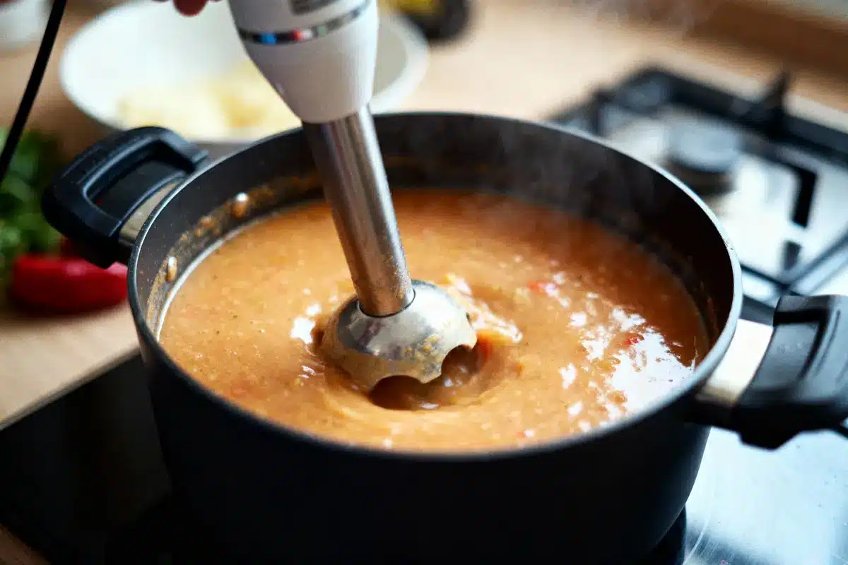 Mixeur plongeant mélange soupe chaude dans une casserole sur une cuisinière, vapeur visible.