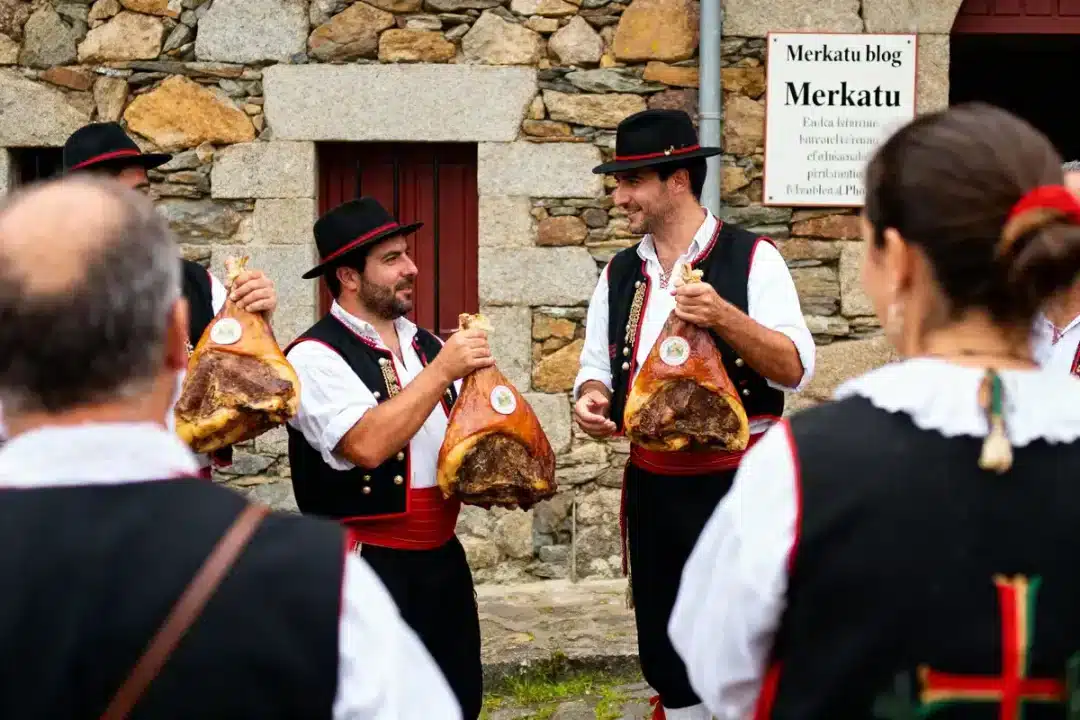 Personnes en costume traditionnel tenant des jambons, discutant devant un mur en pierre. Fête folklorique basque.