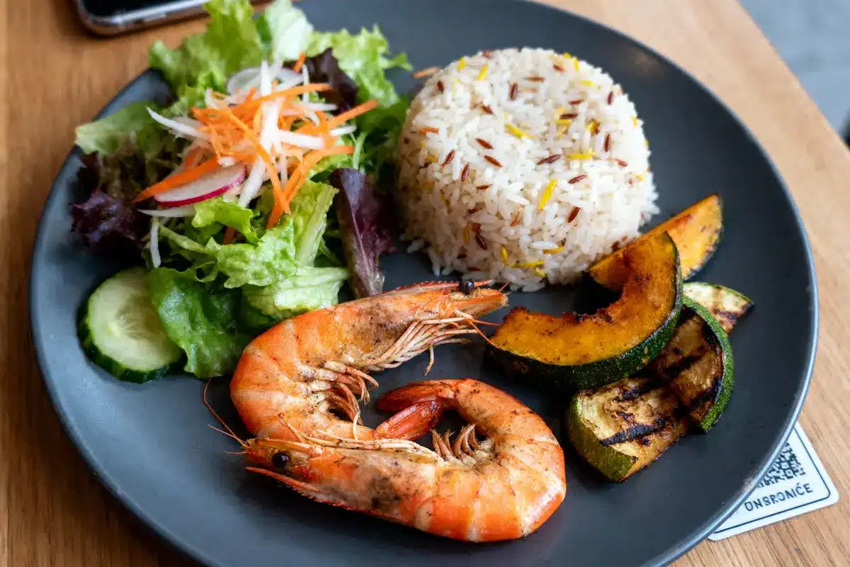 Assiette contenant riz, crevettes grillées, salade verte, légumes grillés et accompagnements variés, sur table en bois.