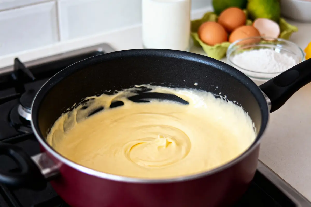 Mélange crémeux dans une poêle, avec œufs, farine et lait en arrière-plan sur le plan de travail.