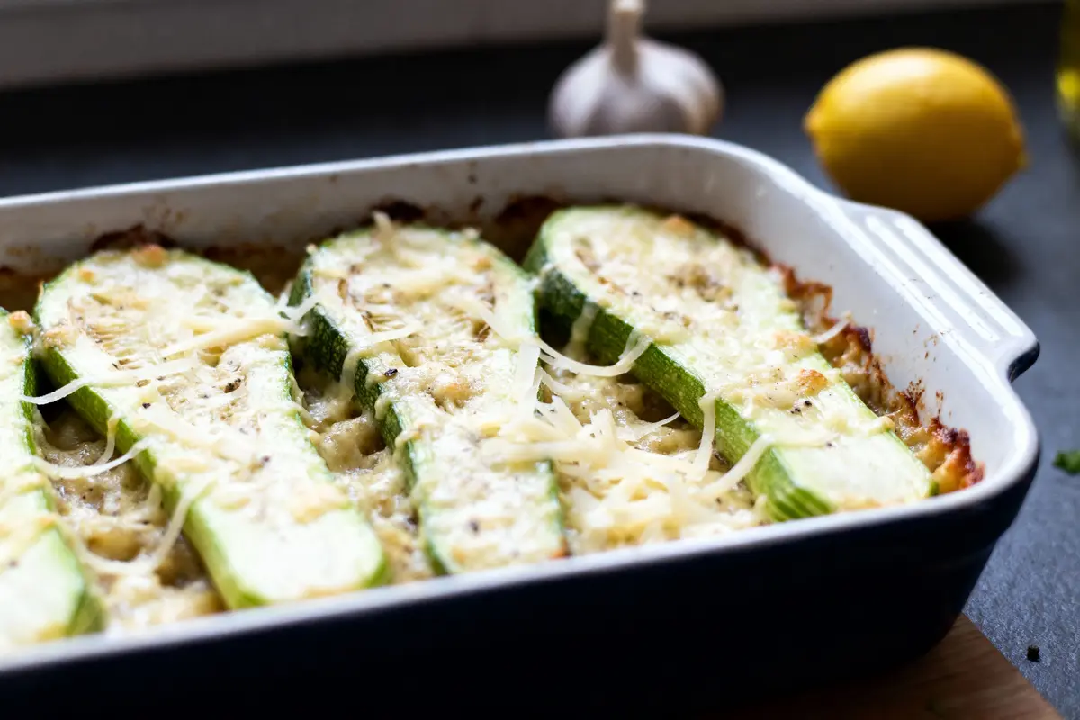 Gratin de courgettes au fromage dans un plat à gratin, avec ail et citron en arrière-plan.