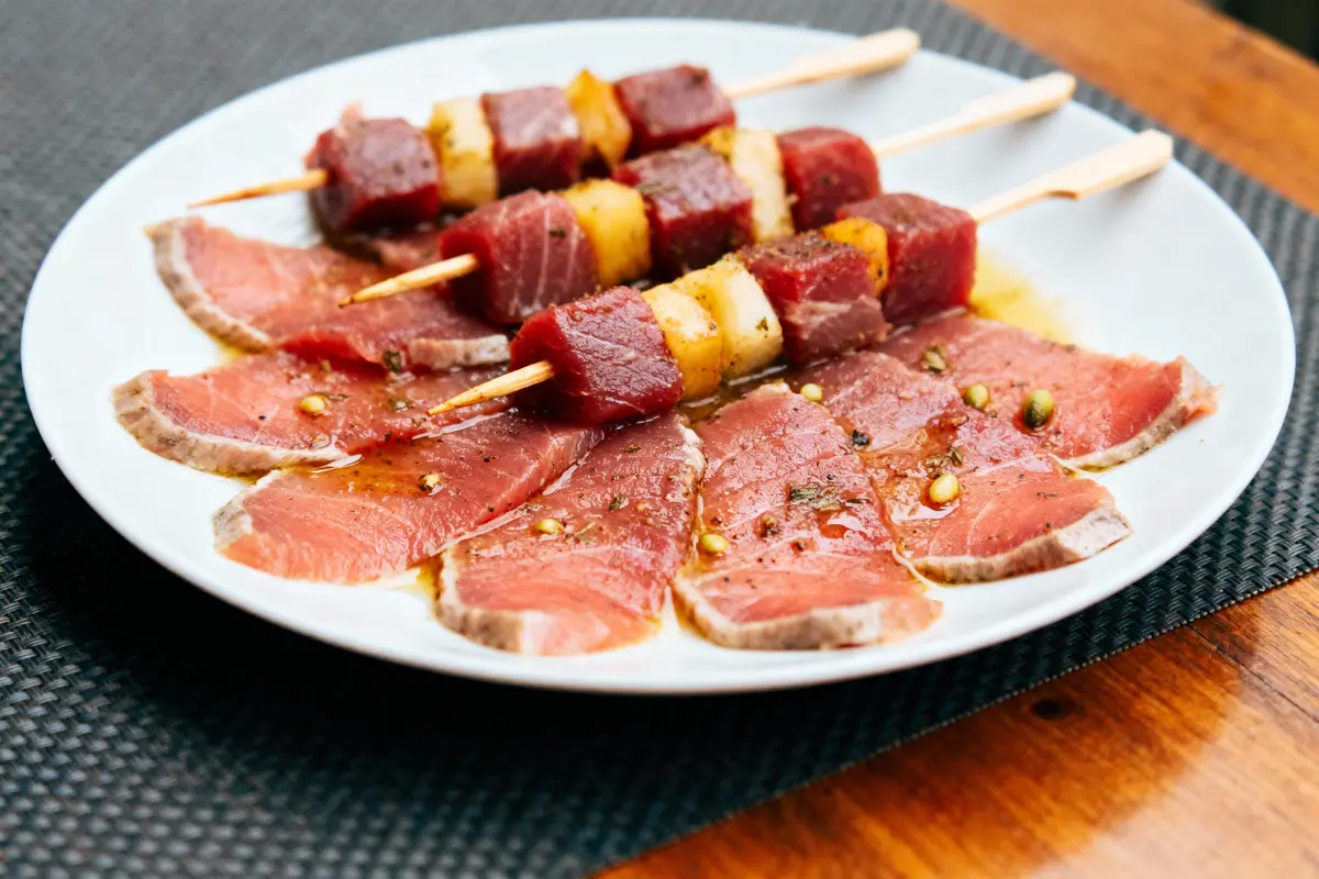 Assiette de carpaccio de thon et brochettes de poisson mariné, servie sur une table en bois.