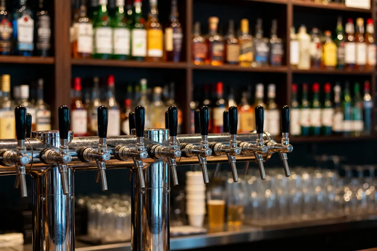 Robinet de bière en métal devant étagères de bouteilles d'alcool et verres, dans un bar.