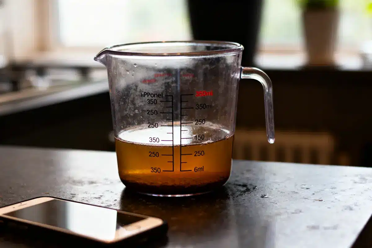 Verre doseur avec liquide brun sur table de cuisine, près d'un smartphone.