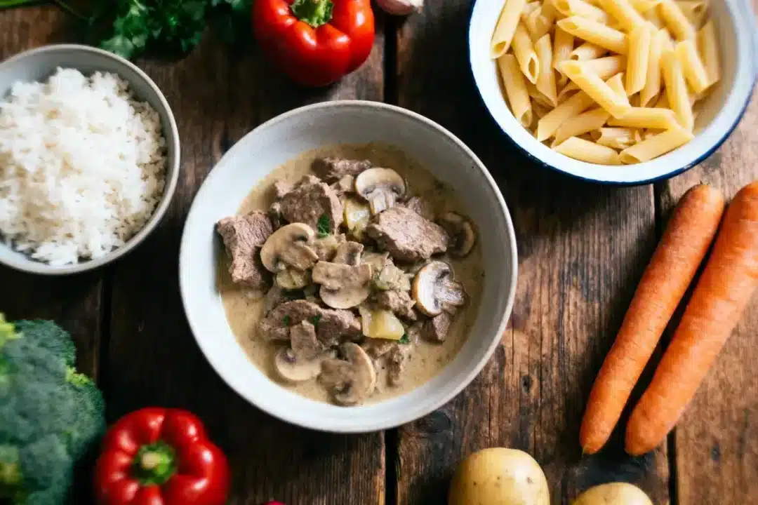 Bol de bœuf aux champignons, avec riz, pâtes, carottes, poivron rouge, brocoli, pommes de terre, sur table bois.