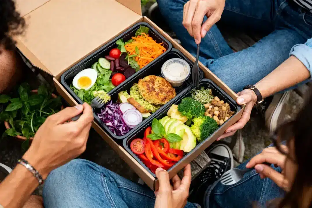 Repas végétarien en boîte, légumes variés, salade, burger végétal, personnes mangeant avec fourchettes noires, ambiance conviviale.