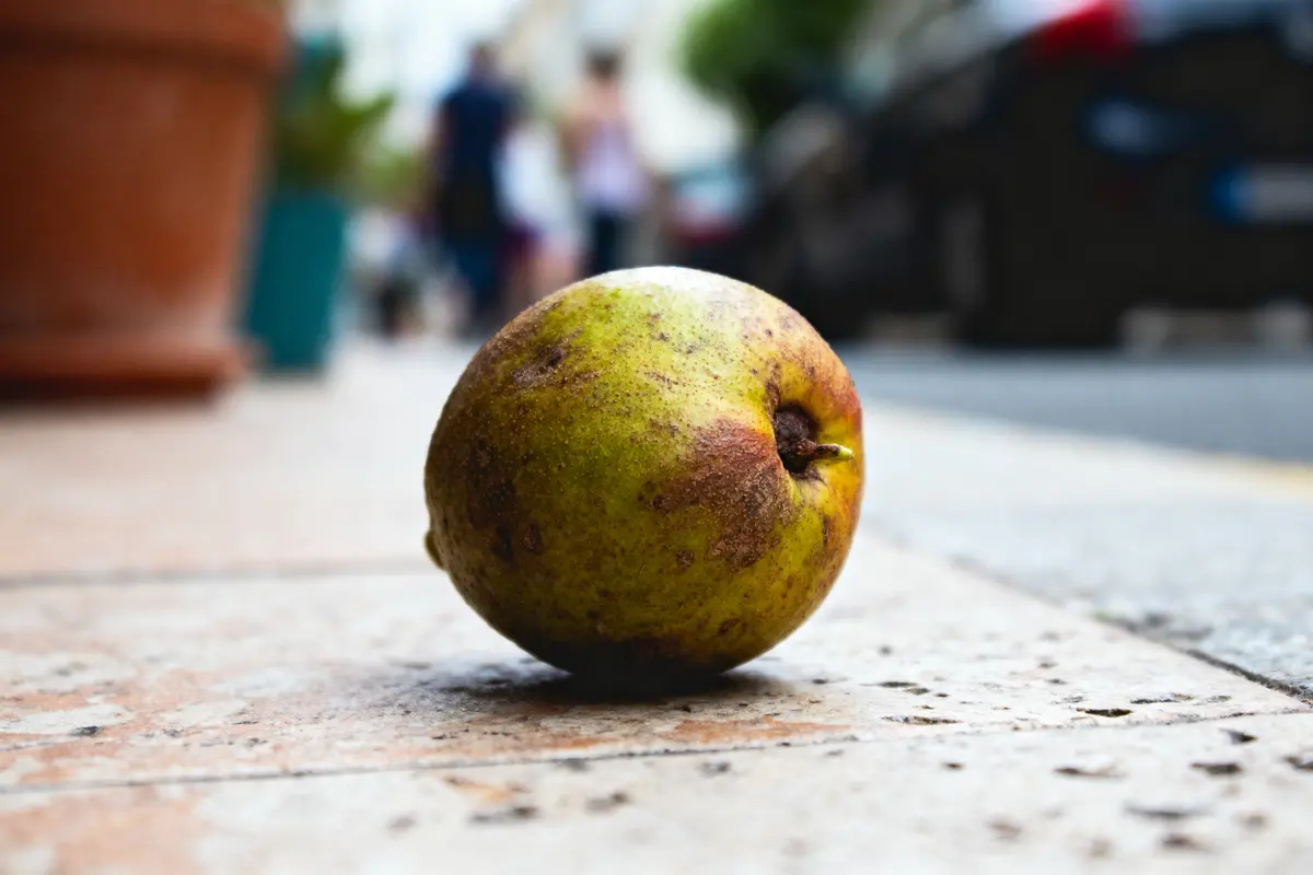 Poire colorée posée sur le trottoir, arrière-plan flou avec passants et pots de fleurs.