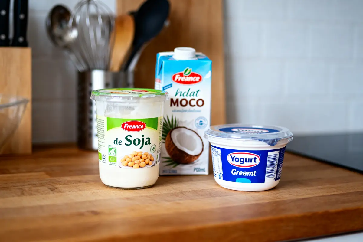 Trois produits alimentaires sur un comptoir de cuisine : yaourt, boisson coco, yaourt soja.