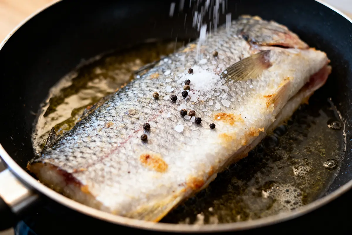 Filet de poisson frit dans une poêle, assaisonné de sel et de grains de poivre.