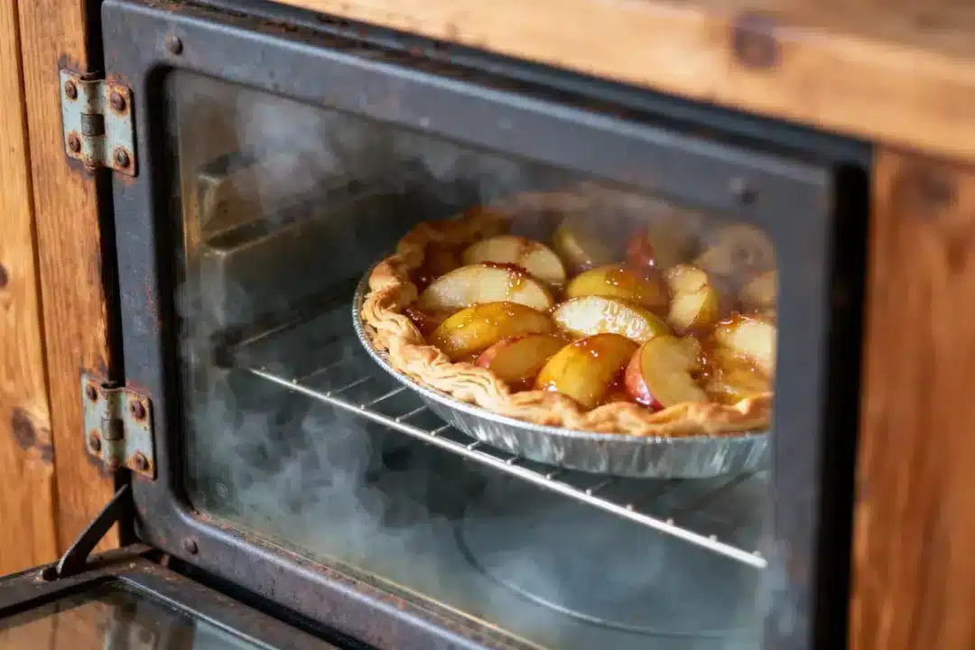 Tarte aux pommes dorée cuisant au four, dans un moule en aluminium, vue à travers la porte en verre.