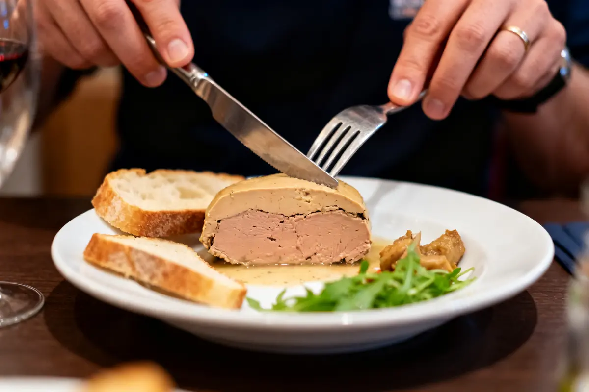 Personne coupant du foie gras avec pain et salade verte sur une assiette blanche